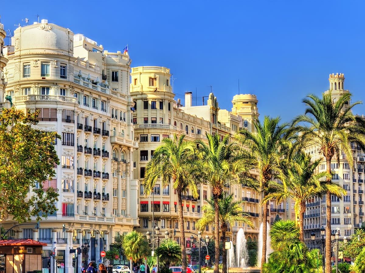 Eine belebte Straße in Valencia mit eleganten historischen Gebäuden, Palmen und einem Brunnen unter blauem Himmel.
