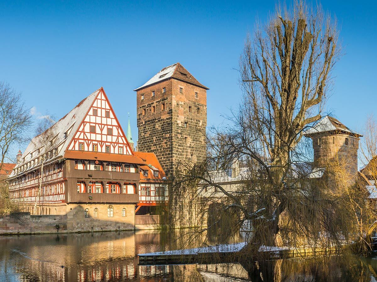 Winterliche Ansicht von Nürnberg mit schneebedecktem Fachwerkhaus, historischen Türmen und einem Fluss unter blauem Himmel.