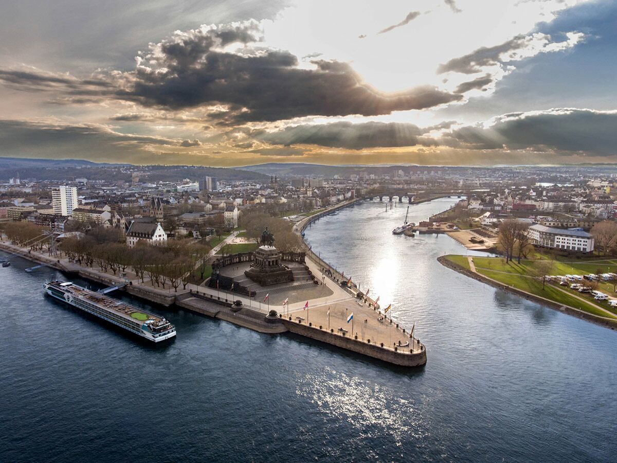 Luftaufnahme von Koblenz am Deutschen Eck mit Rhein und Mosel, historischem Denkmal, Flusskreuzfahrtschiff und Stadtpanorama unter dramatischem Himmel.