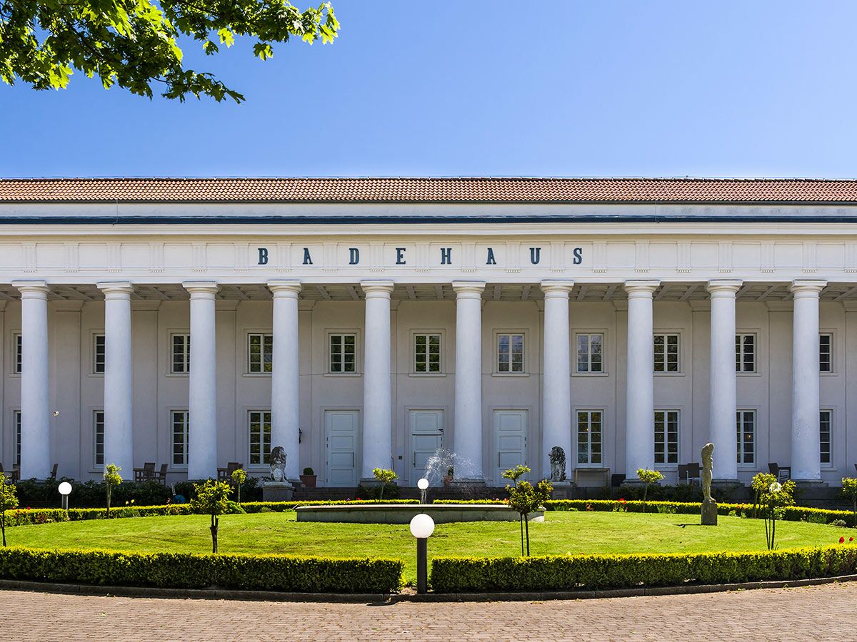 Historisches Badehaus mit weißer Kolonnade und der Aufschrift "BADEHAUS" umgeben von einem grünen Garten unter blauem Himmel.