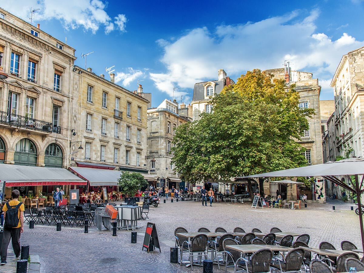 Sonnenbeschienener Stadtplatz in Bordeaux mit historischen Gebäuden, großen Bäumen und zahlreichen Outdoor-Cafés voller Menschen.