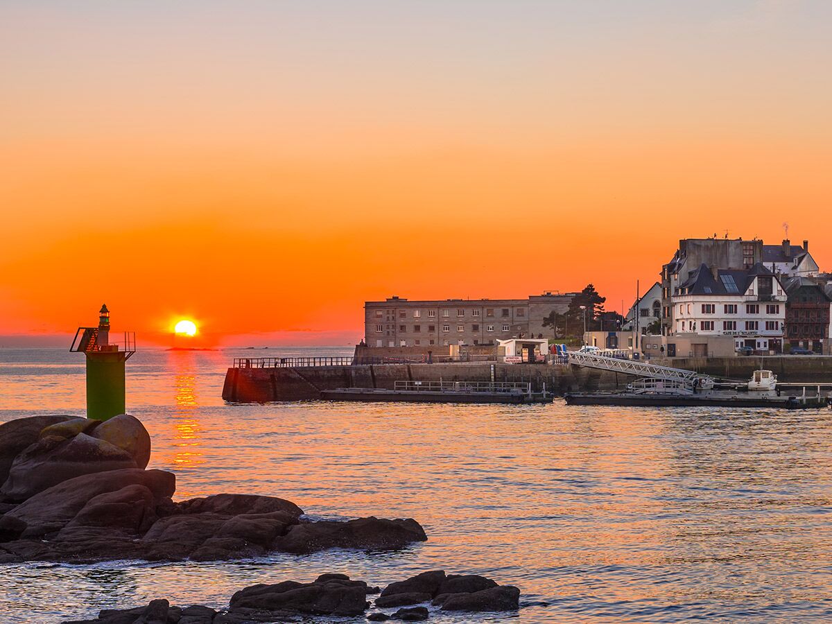 Malerischer Sonnenaufgang über einem Hafen mit grünem Leuchtturm auf Felsen, Gebäuden und goldenem Wasser.