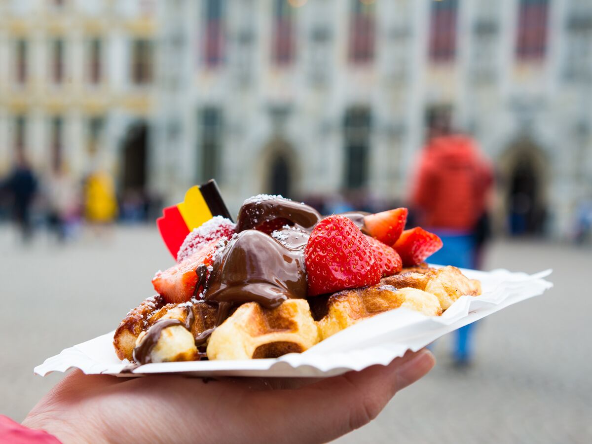 Eine Hand hält belgische Waffeln mit flüssiger Schokolade, frischen Erdbeeren, Puderzucker und einer belgischen Flagge auf Pappteller.