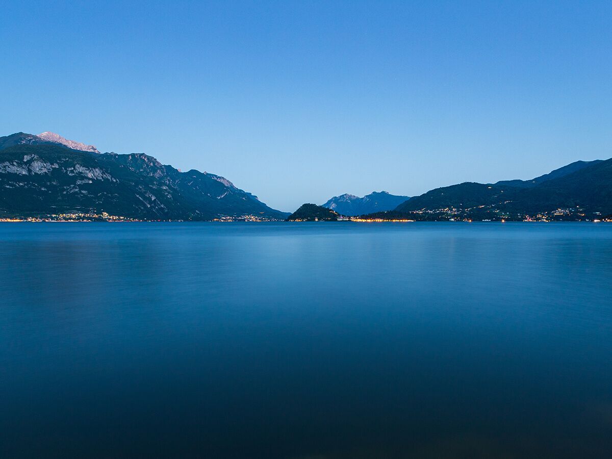 Panoramablick auf einen ruhigen Bergsee bei Abenddämmerung, umgeben von beleuchteten Uferstädten und schroffen Bergen.