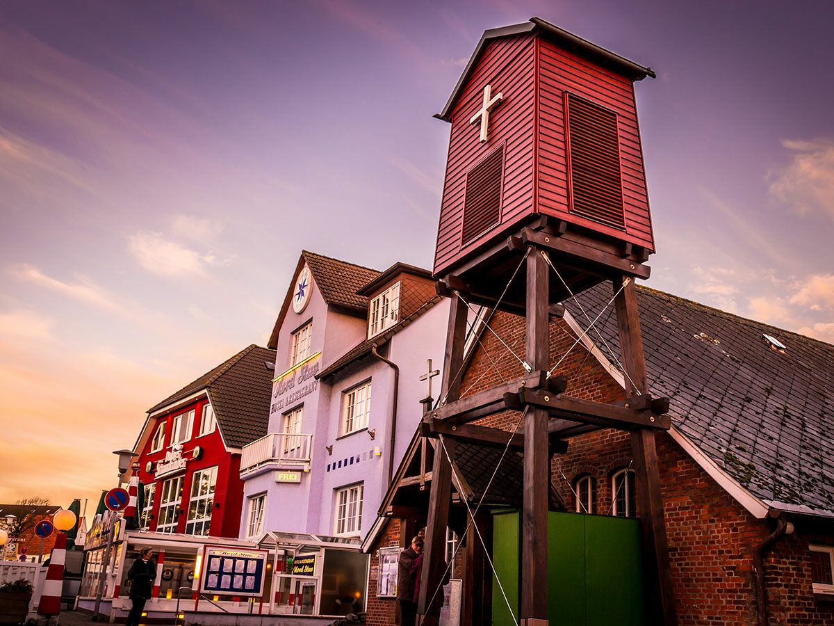Roter hölzerner Glockenturm mit Kreuz, bunte Gebäude und Geschäfte in einer Küstenstadt bei Sonnenuntergang.