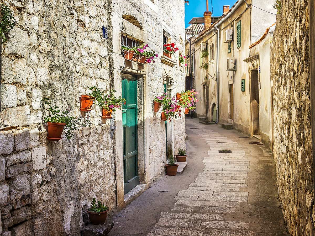 Enge historische Gasse mit groben Steinmauern, grüner Tür und vielen Blumentöpfen voller bunter Pflanzen in mediterraner Stadt.