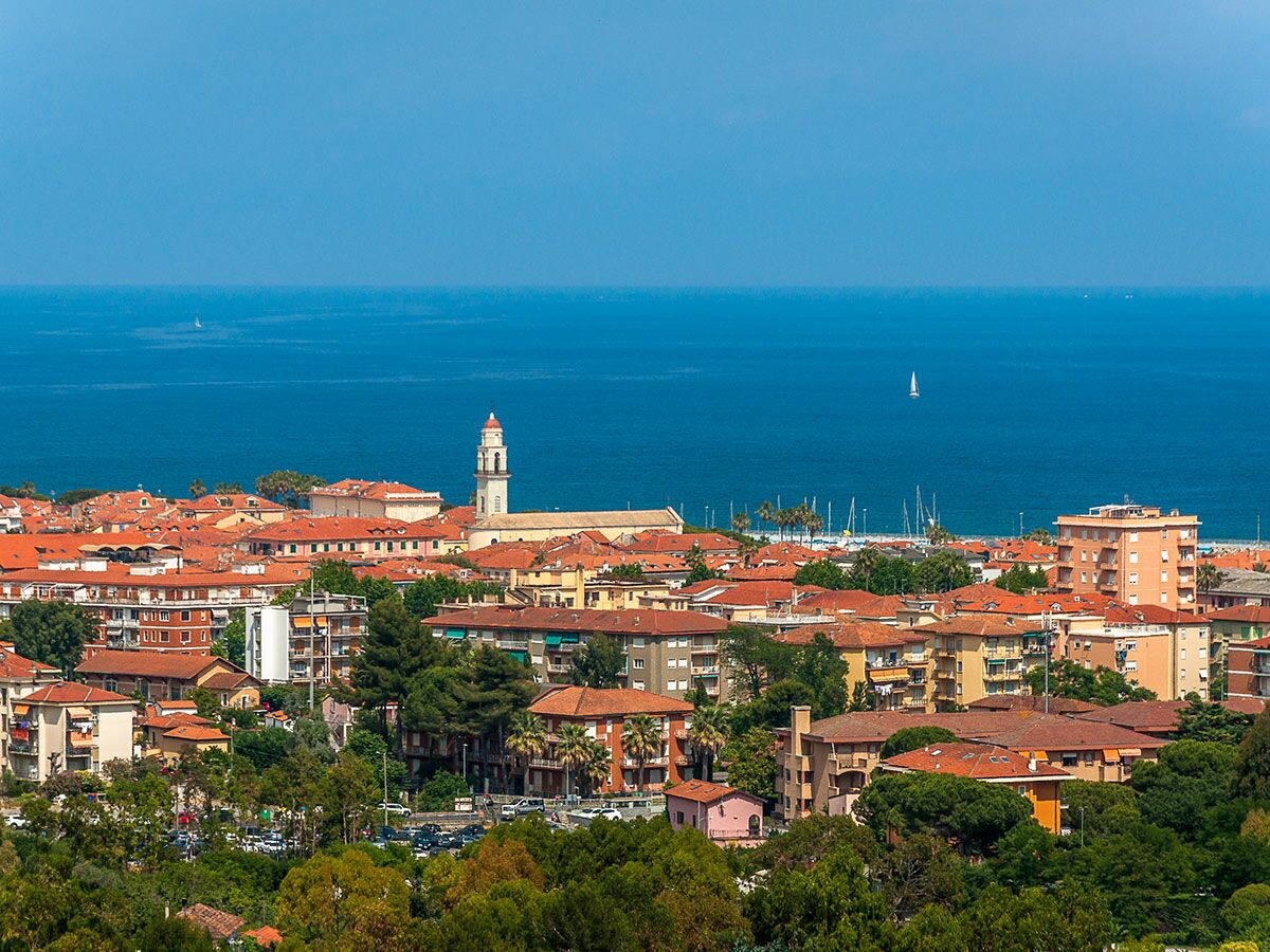 Mittelmeer-Küstenstadt mit roten Dächern, Kirchturm, grünen Bäumen, blauem Meer und Segelbooten unter klarem Himmel.