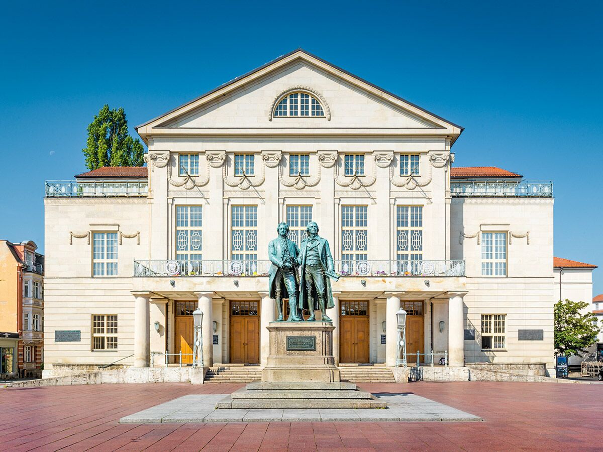 Das Deutsche Nationaltheater in Weimar mit dem Goethe-Schiller-Denkmal bei strahlendem Sonnenschein. Historisches Gebäude und Statuen.