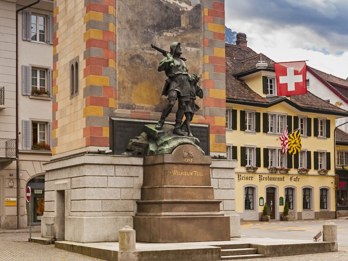 Bronzestatue von Wilhelm Tell und seinem Sohn vor bemalter Säule in Altdorf, Schweiz, mit Schweizer Flagge.