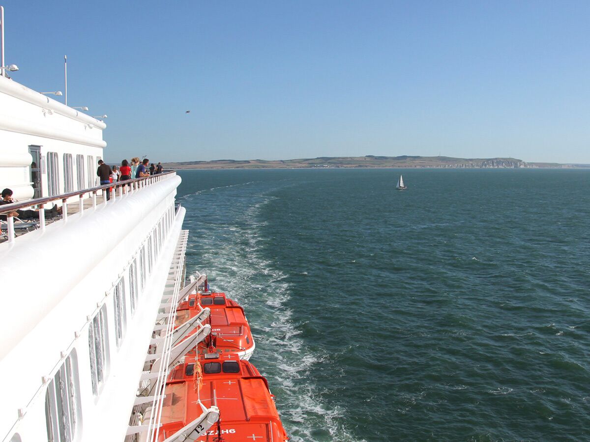 Passagiere auf dem Deck einer weißen Fähre blicken über das Meer, vorbei an orangefarbenen Rettungsbooten, auf eine ferne Küste und ein Segelboot.