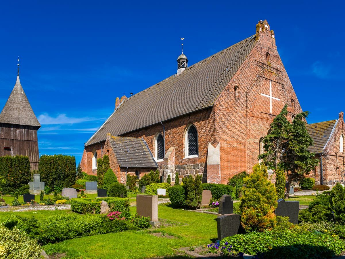 Historische Backsteinkirche mit separatem Glockenturm auf grünem Friedhof, sonniger Tag mit blauem Himmel.
