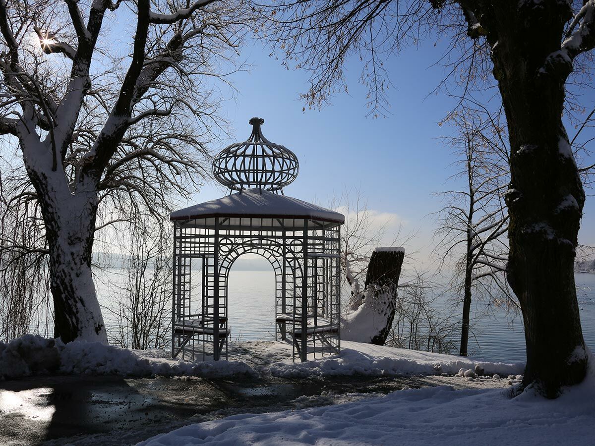 Verschneiter Metallpavillon am ruhigen Seeufer mit großen Bäumen, unter blauem Himmel und Sonnenschein.
