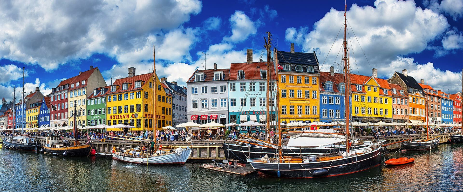 Panoramaansicht des Nyhavn in Kopenhagen mit bunten Gebäuden, Segelbooten und vielen Menschen am Kai unter blauem Himmel.
