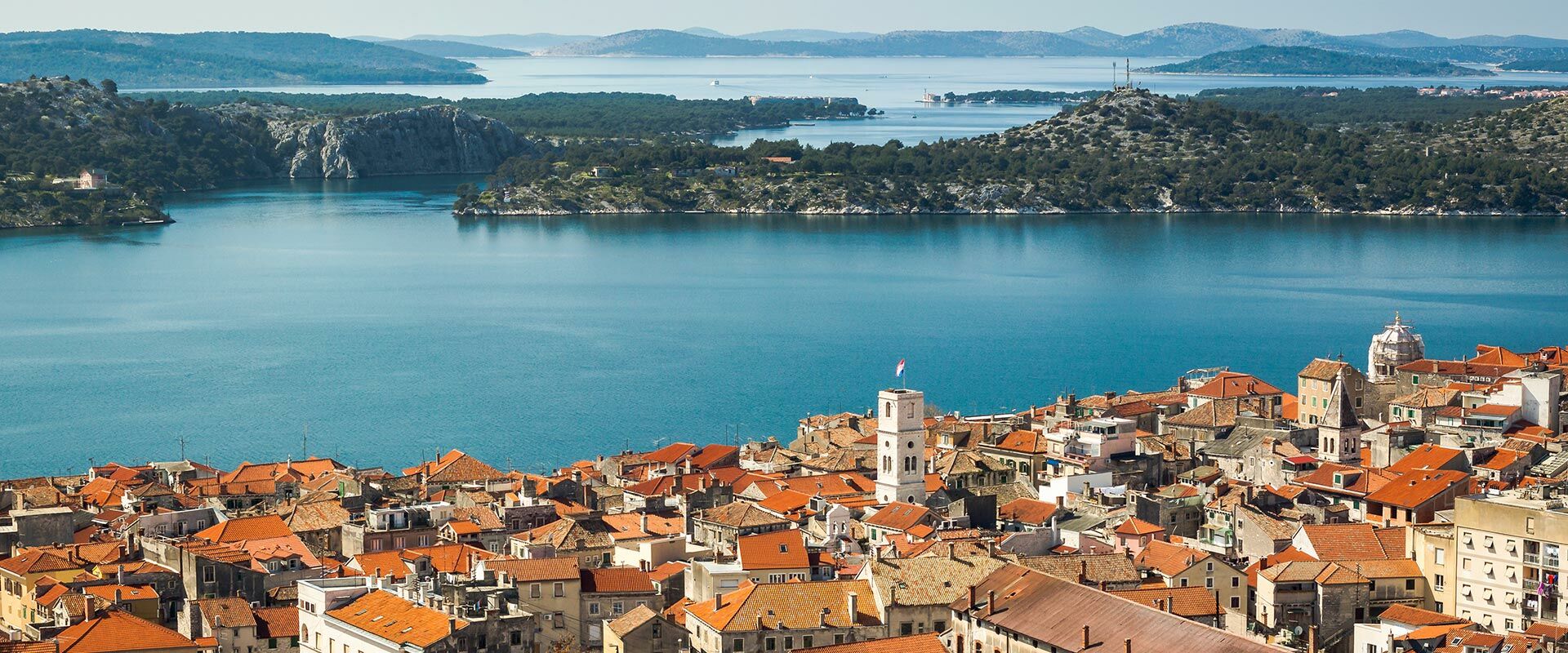 Panoramablick auf die historische kroatische Stadt Šibenik mit roten Dächern, Kirche und blauer Adria im Hintergrund, umgeben von grünen Hügeln.