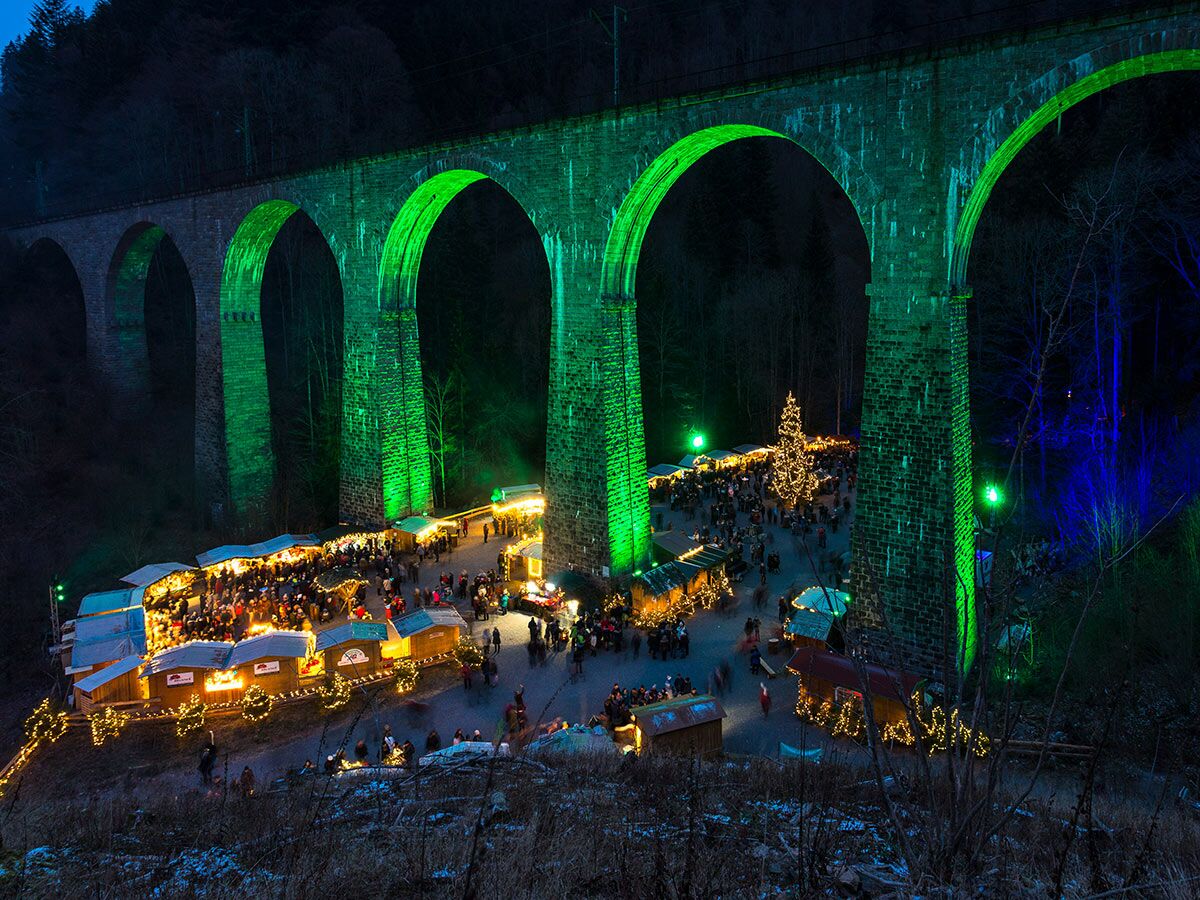 Festlicher Weihnachtsmarkt mit zahlreichen Besuchern unter einem imposanten, grün beleuchteten Steinviadukt bei Nacht.
