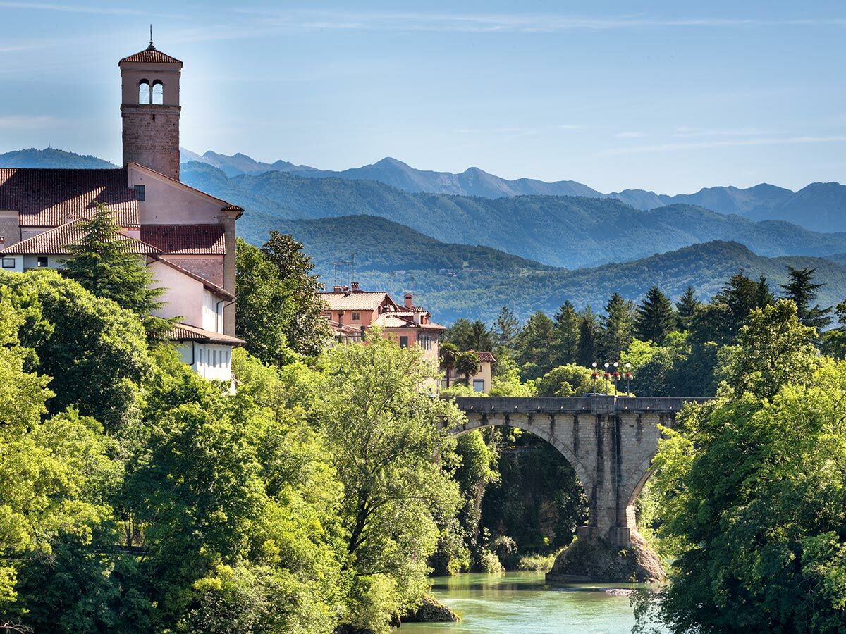 Historische Stadt mit Kirchturm, roter Dächer, Steinbrücke über Fluss, umgeben von grünen Bäumen und Bergen unter blauem Himmel.