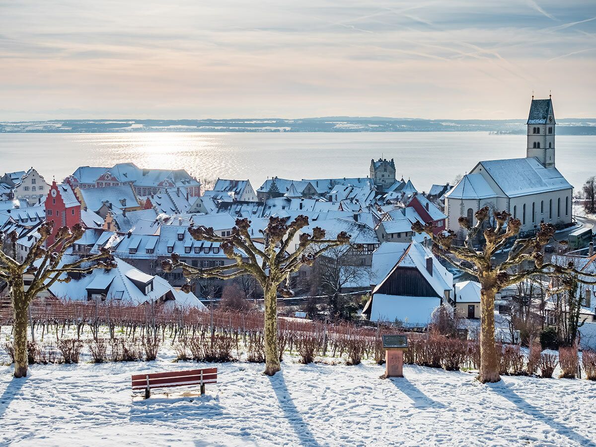 Winterlandschaft mit verschneiter europäischer Stadt am See, Kirchturm, alten Häusern und spiegelndem Wasser.