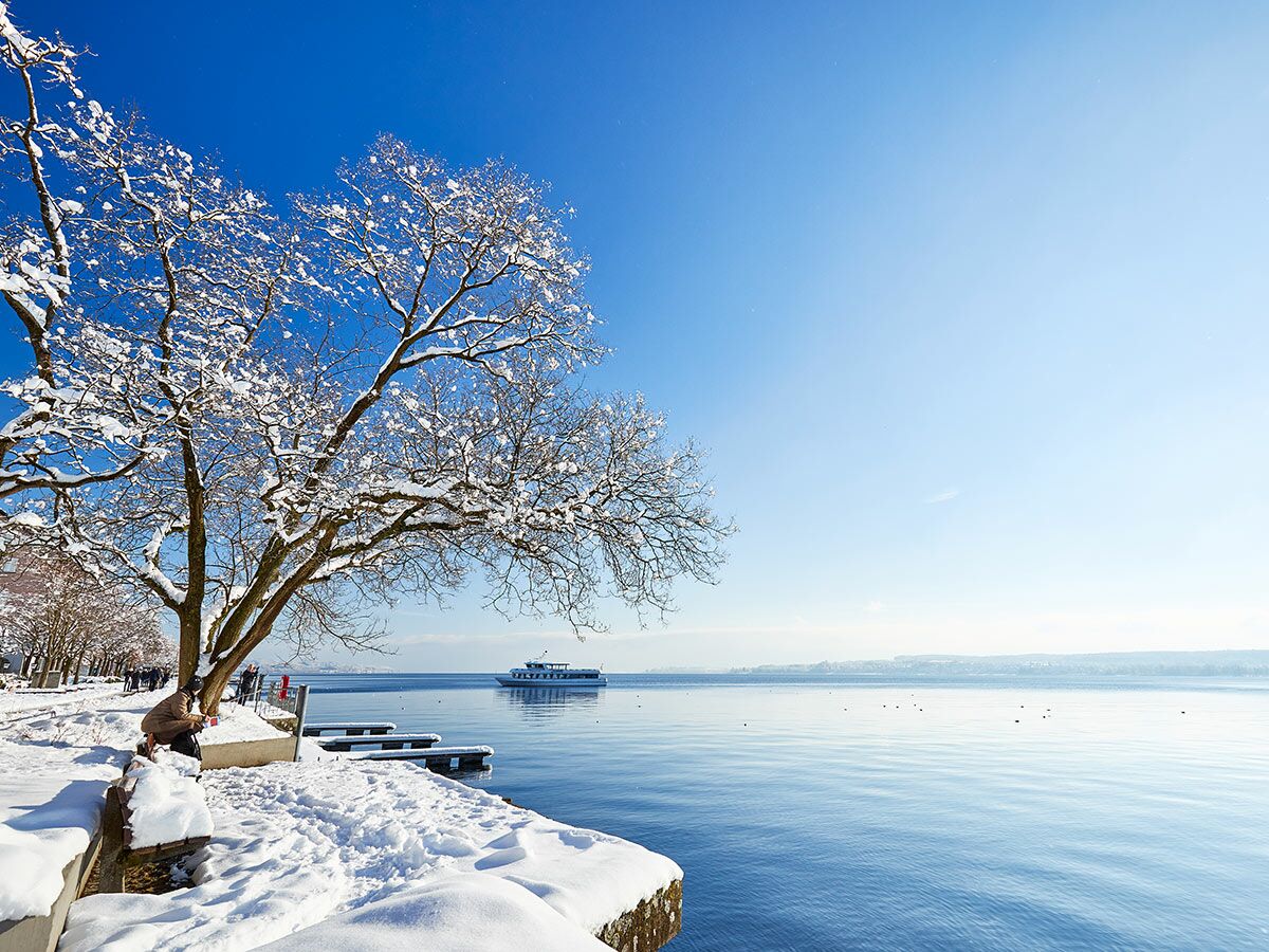 Idyllische Winterlandschaft am Seeufer mit verschneiten Bäumen, Menschen und einem Schiff auf blauem Wasser unter klarem Himmel.