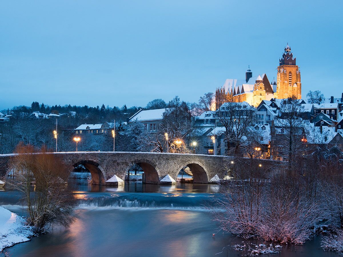 Panoramablick auf eine verschneite historische Stadt am Abend mit einer beleuchteten Kirche und alter Steinbrücke über einen Fluss.