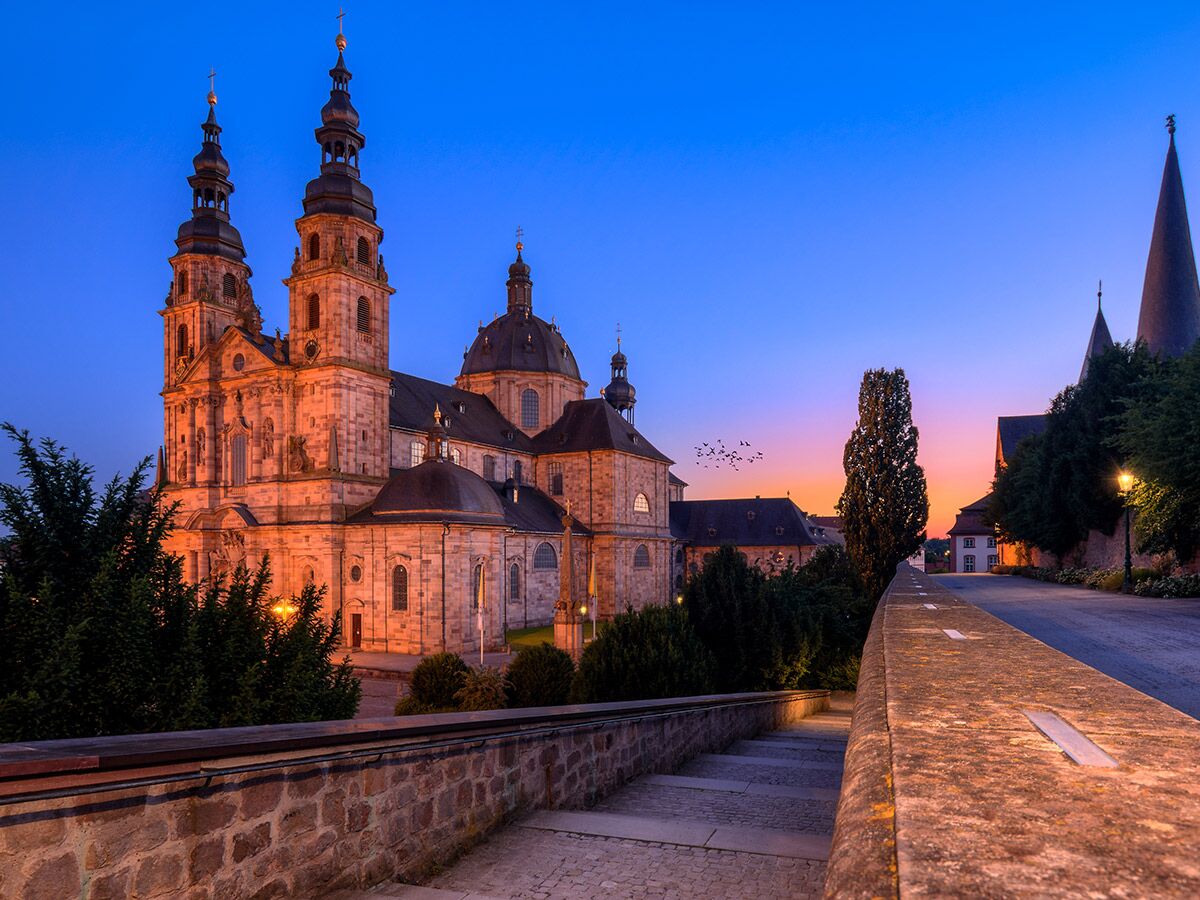Barocke Kathedrale in Fulda bei Dämmerung mit leuchtenden Fenstern, farbigem Himmel, Bäumen und einer Mauer.