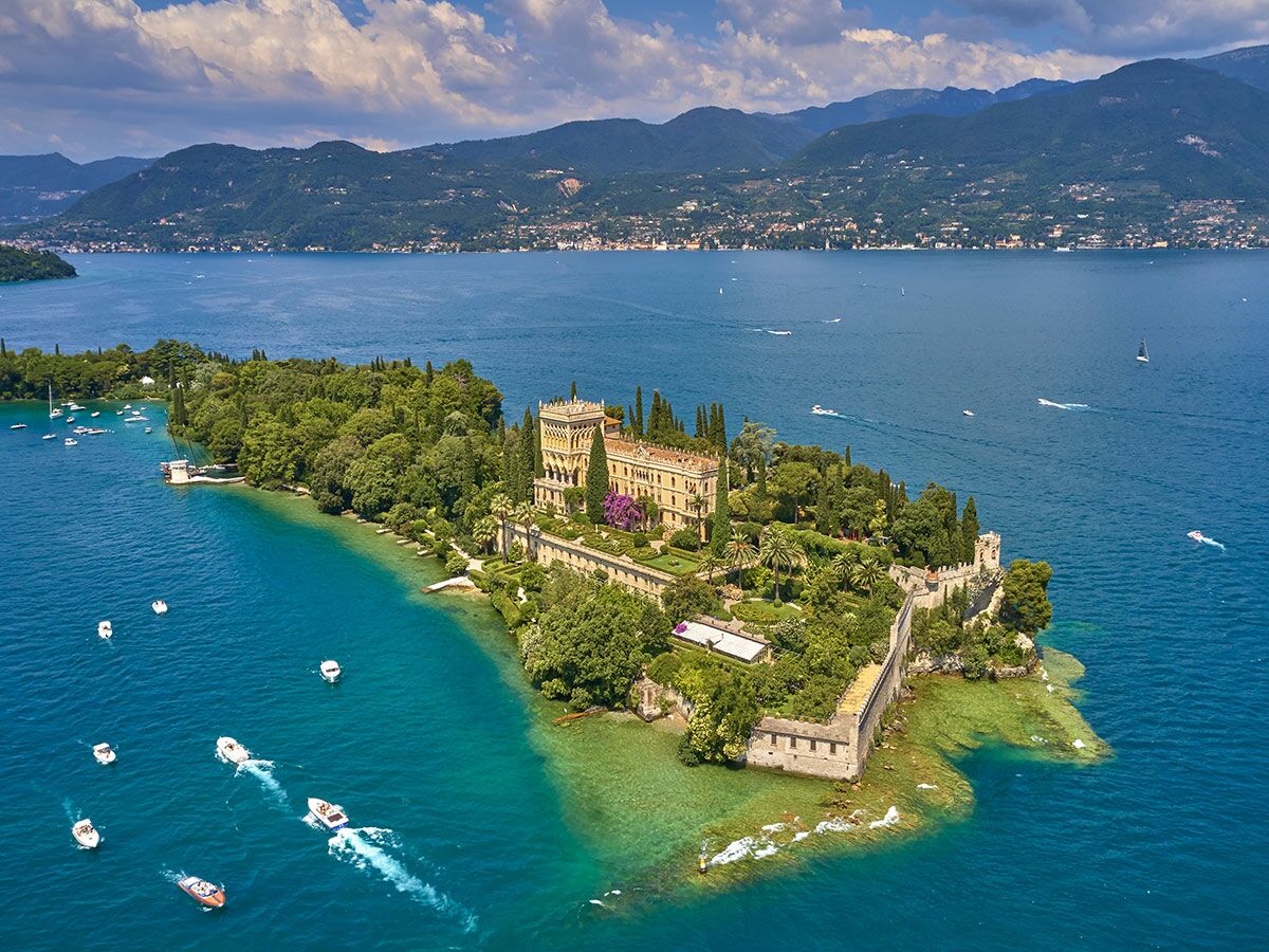 Malerische Isola del Garda mit historischer Villa, üppigem Garten und Booten im türkisfarbenen Wasser des Gardasees vor Bergkulisse.