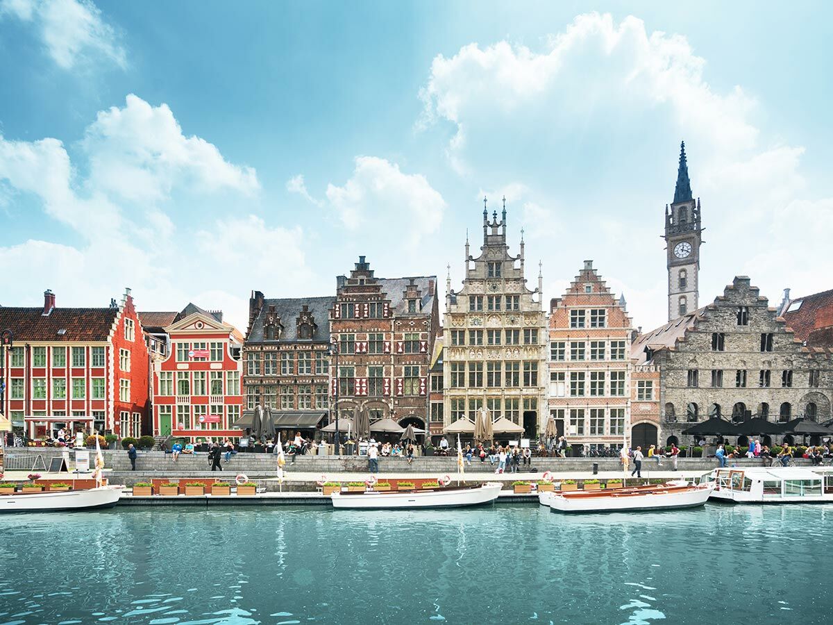 Blick auf historische Giebelhäuser und einen Kanal in Gent, Belgien, mit Menschen am Ufer und Booten im Wasser.