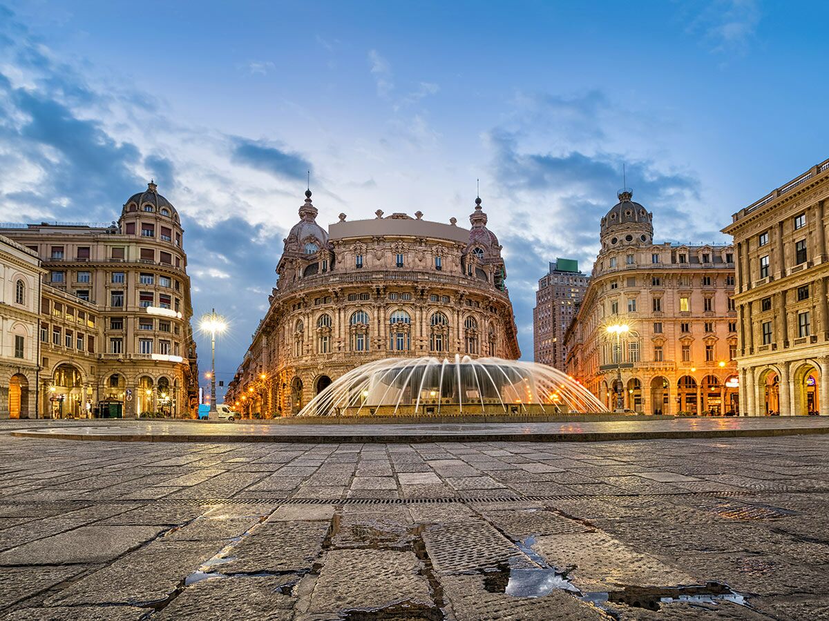 Piazza De Ferrari in Genua bei Dämmerung mit leuchtenden historischen Gebäuden, beleuchtetem Brunnen und nassem Kopfsteinpflaster.
