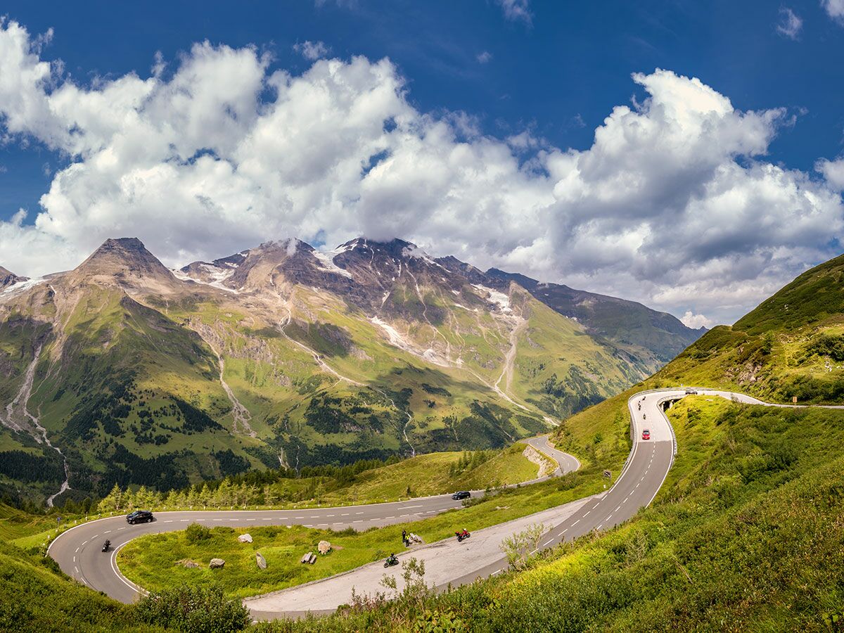 Eine kurvenreiche Alpenstraße schlängelt sich durch eine grüne Berglandschaft mit schneebedeckten Gipfeln unter blauem Himmel.