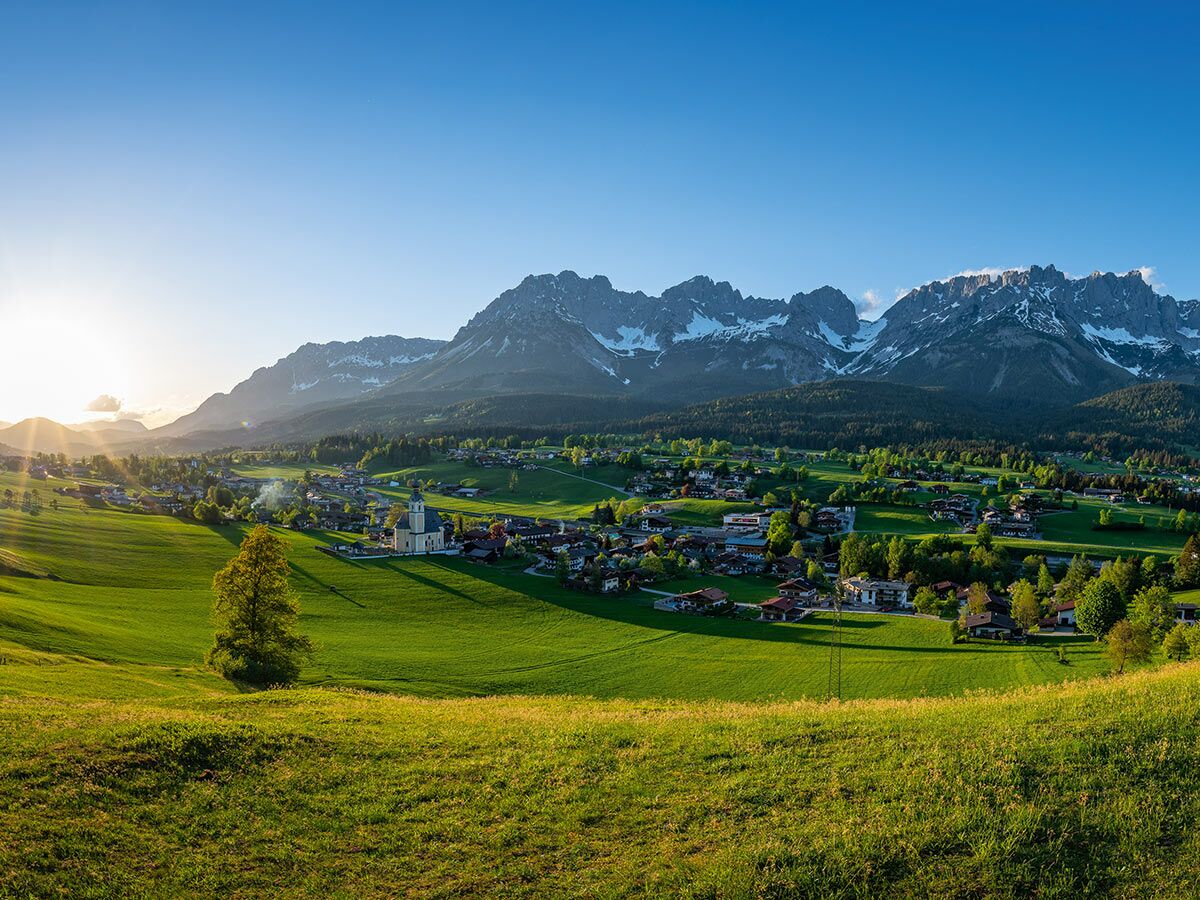 Idyllisches Alpendorf am Wilden Kaiser bei Sonnenuntergang, mit Kirche, grünen Wiesen und schneebedeckten Bergen.
