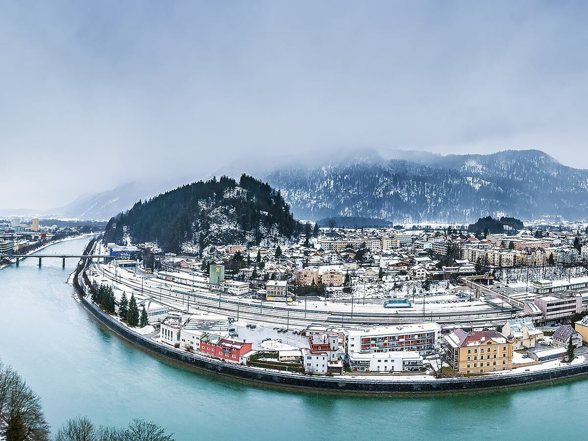 Panoramablick auf eine schneebedeckte Alpenstadt mit einem türkisfarbenen Fluss und Eisenbahnstrecken vor bewaldeten Bergen.