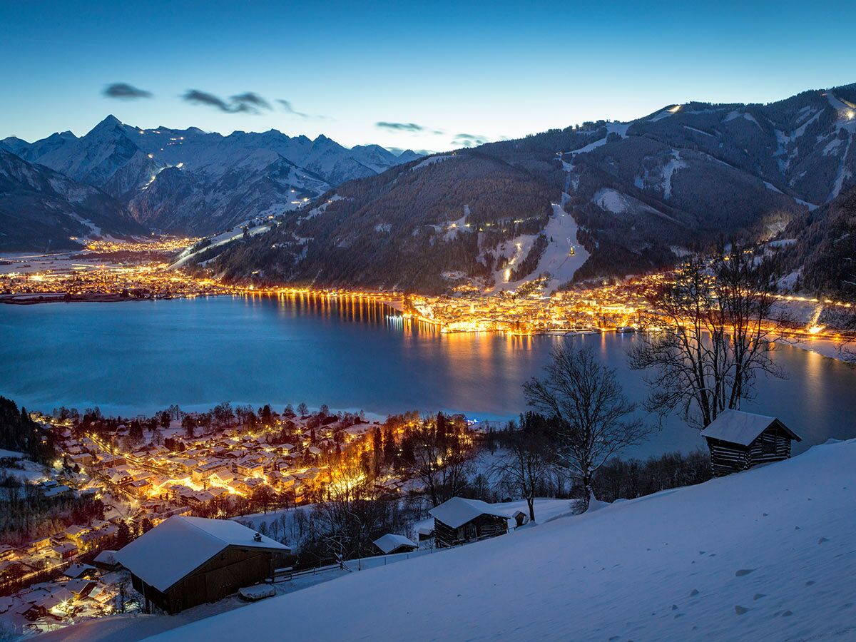 Winterlicher Panoramablick auf das beleuchtete Zell am See am Seeufer, umgeben von schneebedeckten Bergen und Skihängen.