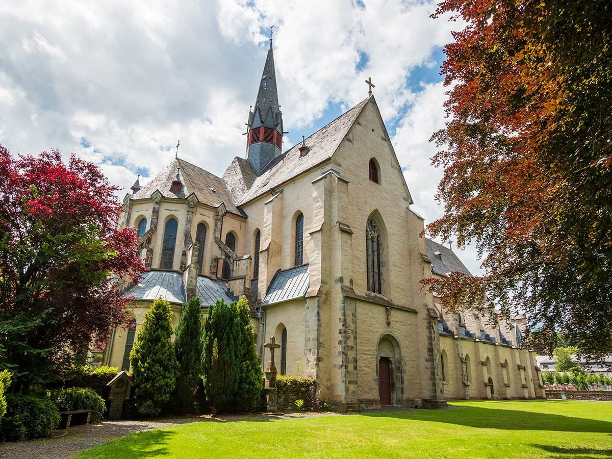 Historische Abtei mit hohem Turm, ockerfarbenen Steinmauern und bunten Herbstbäumen in einem grünen Garten unter blauem Himmel.