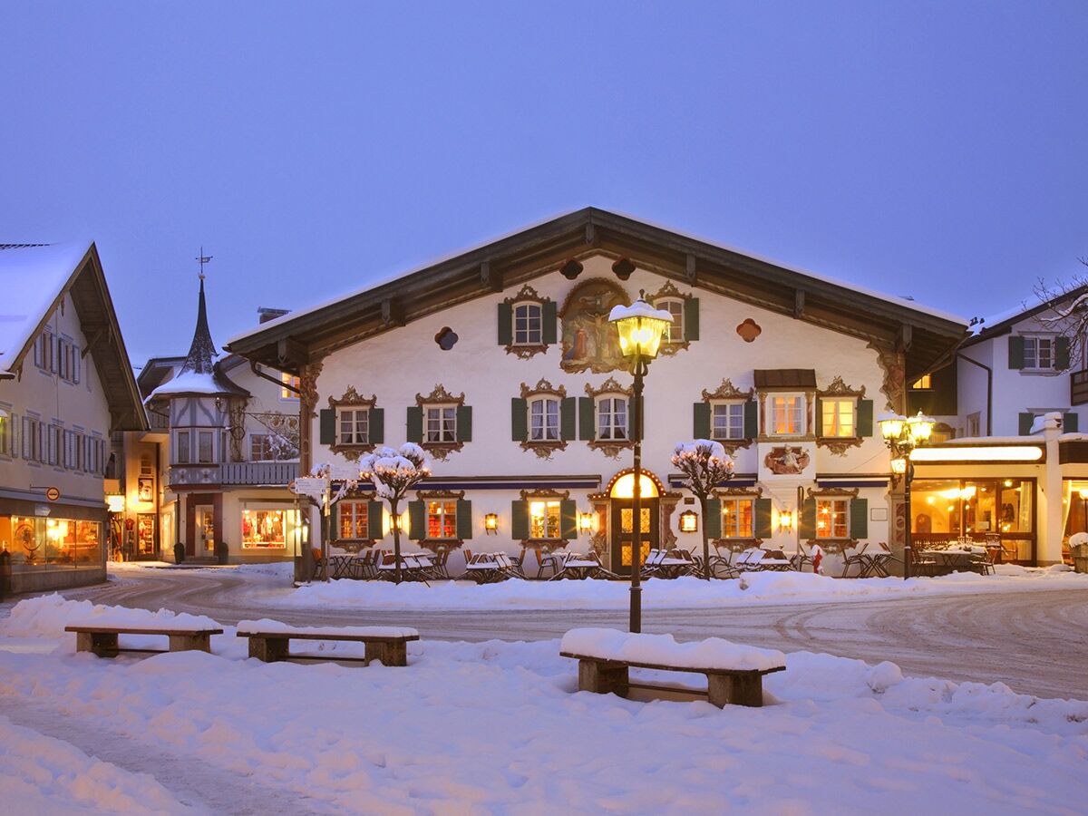 Verschneiter Dorfplatz abends mit beleuchteten, traditionellen Häusern, Giebelfassaden, Geschäften und Bänken unter blauem Himmel.