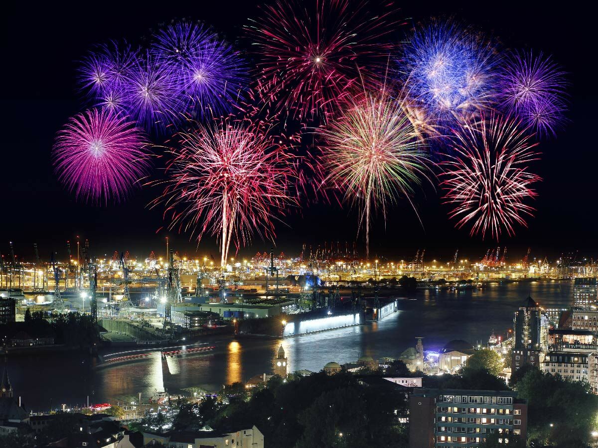 Spektakuläres Feuerwerk über dem Hamburger Hafen bei Nacht, mit bunten Explosionen und beleuchteten Kränen am Wasser.