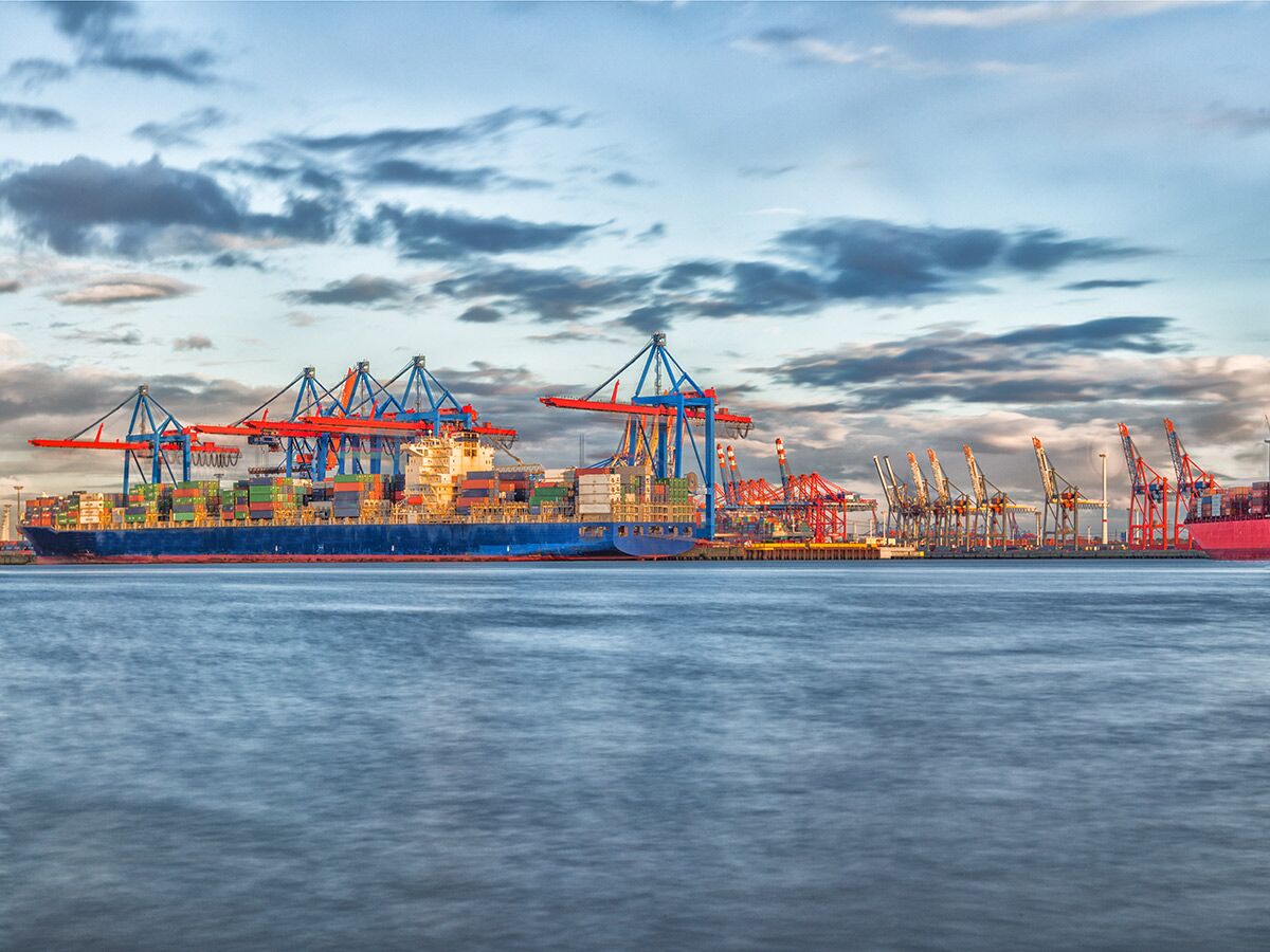 Großes Containerschiff mit vielen Frachtcontainern und roten, blauen Hafenkränen bei Sonnenuntergang im Hamburger Hafen.