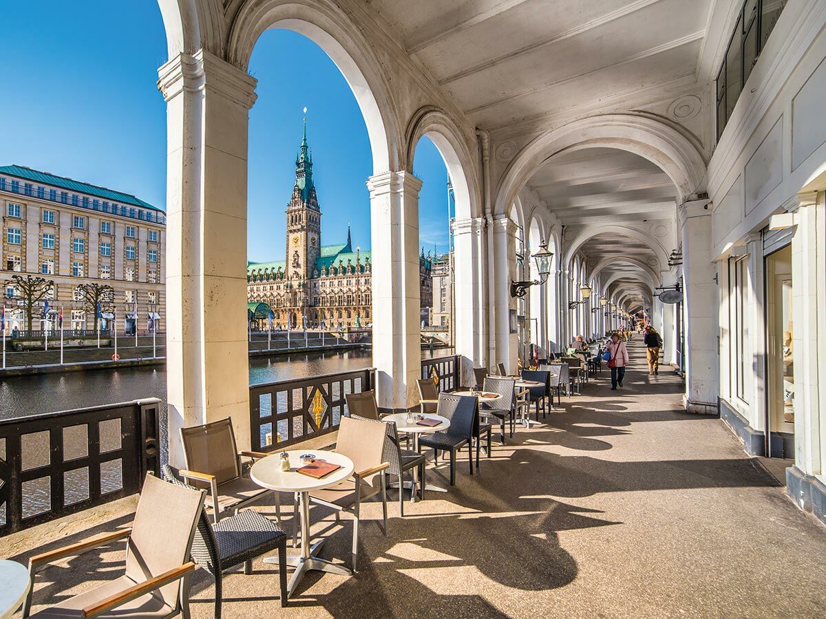 Blick auf Hamburgs Alsterarkaden mit Cafés am Kanal und dem historischen Rathaus im Hintergrund bei Sonnenschein.