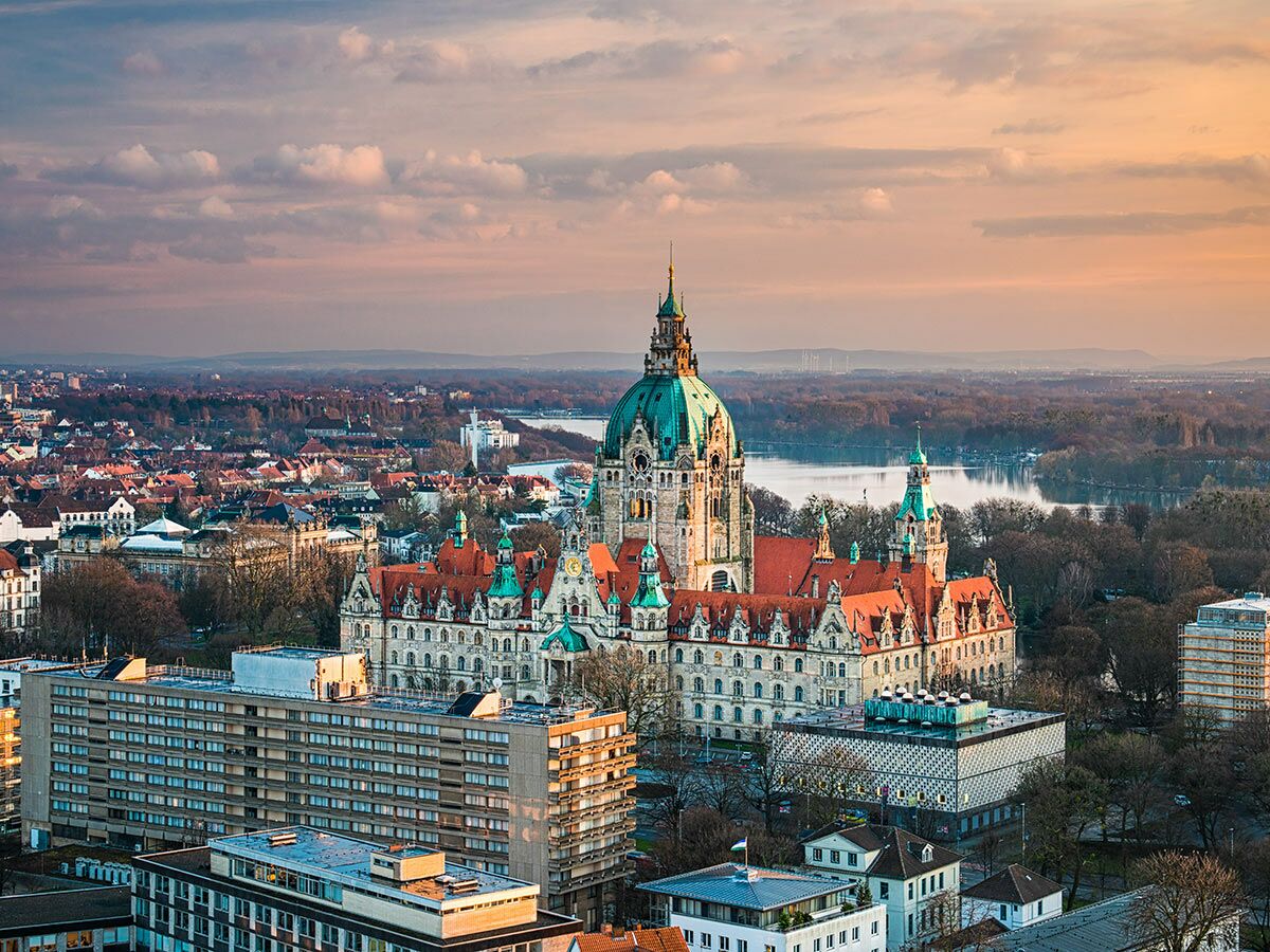 Luftbild des Neuen Rathauses in Hannover mit grünem Dom, umgeben von der Stadtlandschaft und einem See bei Sonnenuntergang.