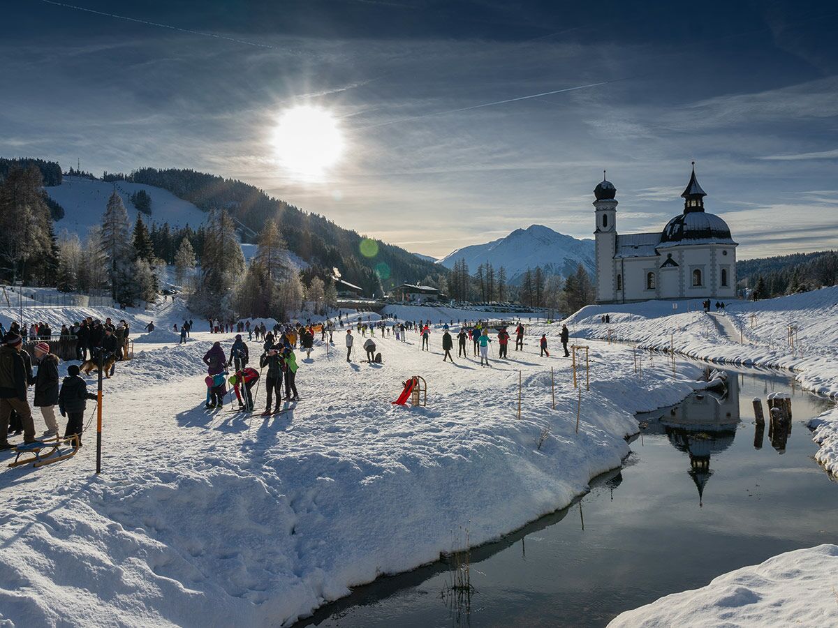 Sonnige Winterlandschaft mit barocker Kirche, spiegelndem Fluss, Bergen und Menschen beim Langlauf in den verschneiten Alpen.