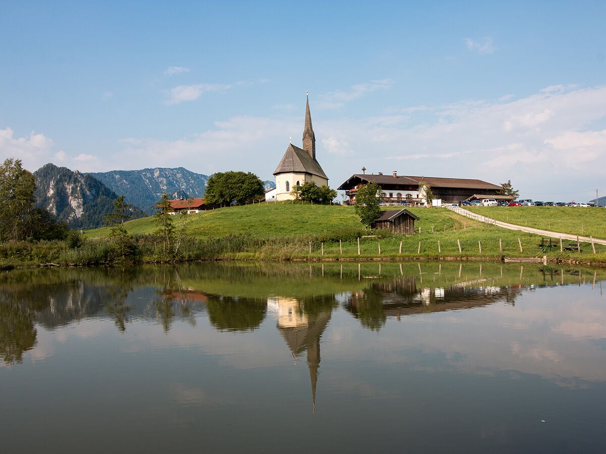 Malerische Alpenkirche am Seeufer mit hohem Kirchturm, umgeben von grünen Hügeln und majestätischen Bergen unter blauem Himmel.