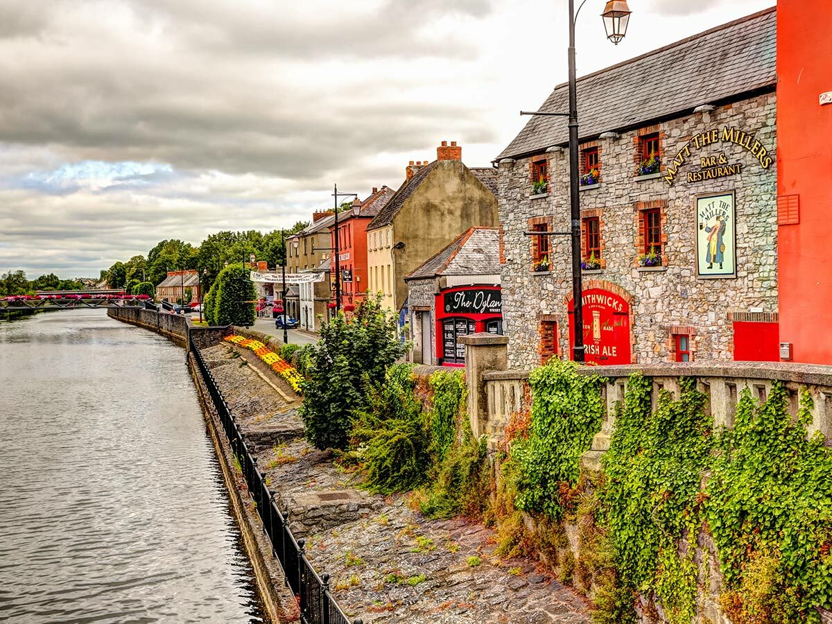 Malerische Steingebäude, Matt The Millers Pub und Flussufer mit Efeu in Kilkenny, Irland, unter bewölktem Himmel.