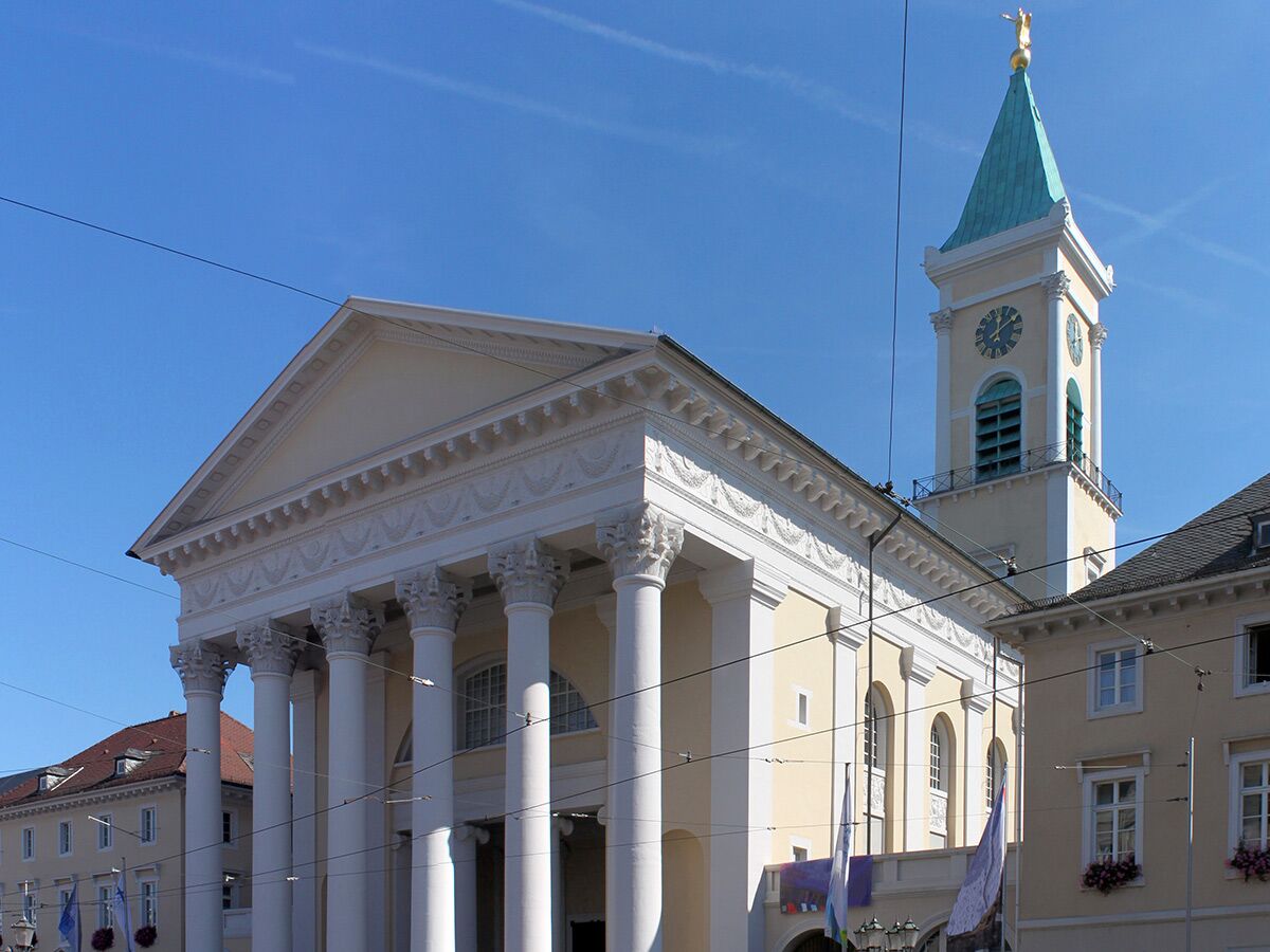 Außenansicht eines großen, neoklassizistischen Gebäudes mit Säulen und Giebel, daneben ein Kirchturm mit grüner Spitze und Uhr unter blauem Himmel.