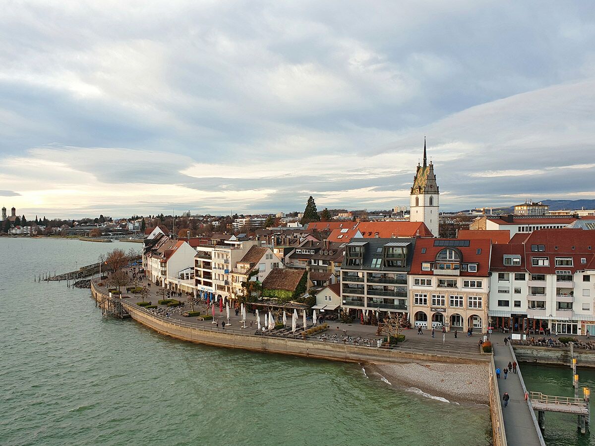 Panorama Konstanz am Bodensee: Uferpromenade, Altstadt, Konstanzer Münster, Hafen, Menschen, Gebäude unter bewölktem Himmel.