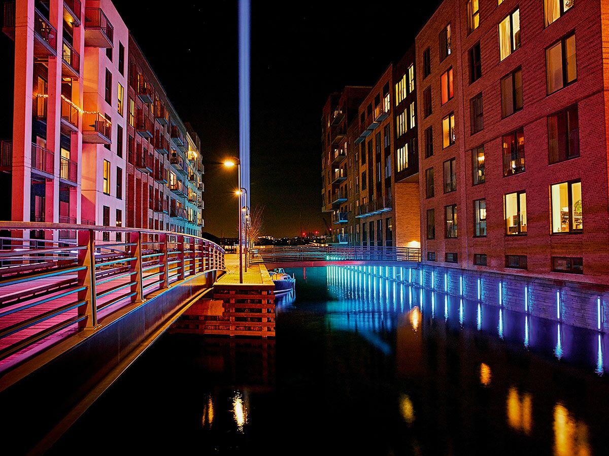 Moderne Wohngebäude und Brücke am beleuchteten Kanal bei Nacht mit rotem und blauem Licht sowie Himmelsstrahl.