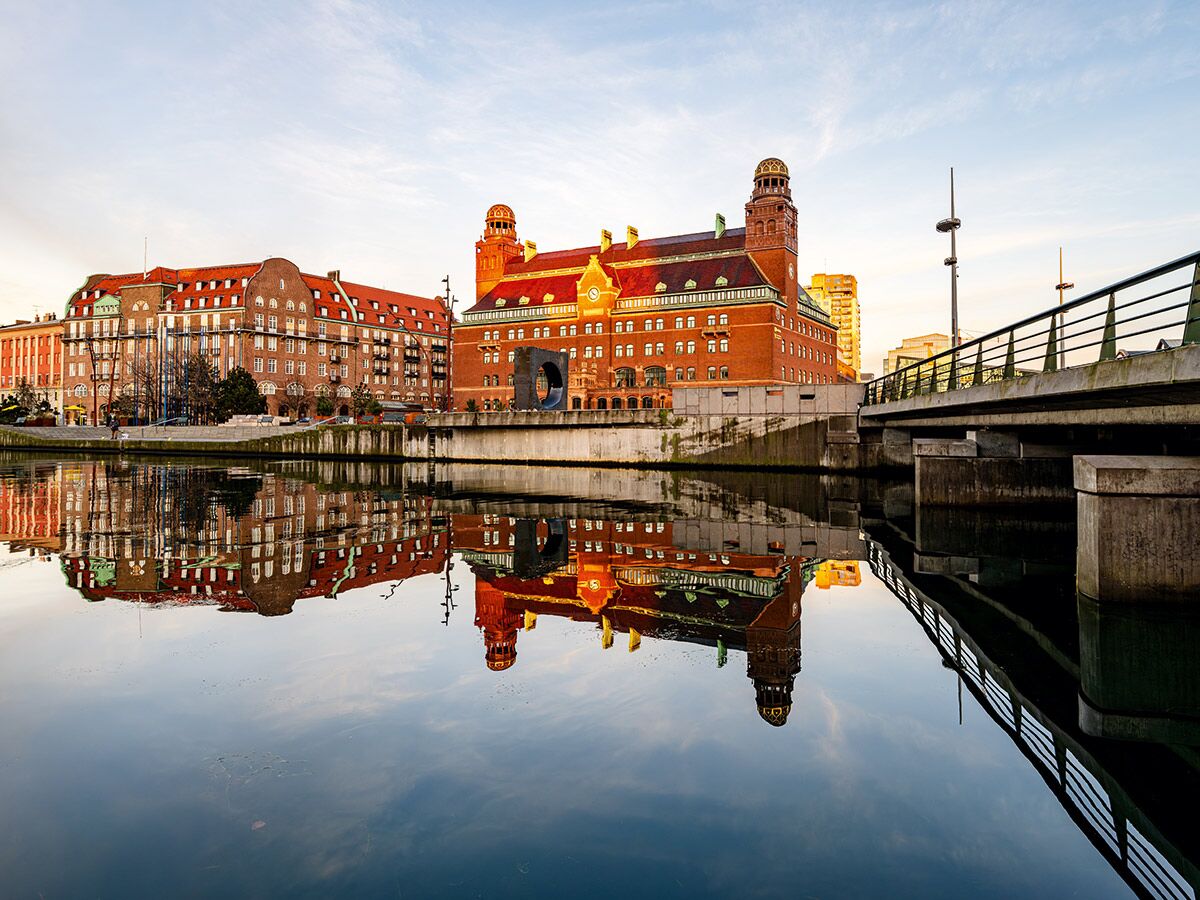 Panoramablick auf rote Backsteingebäude, darunter ein markantes Gebäude mit Türmen, die sich in einem ruhigen Fluss in Malmö spiegeln.