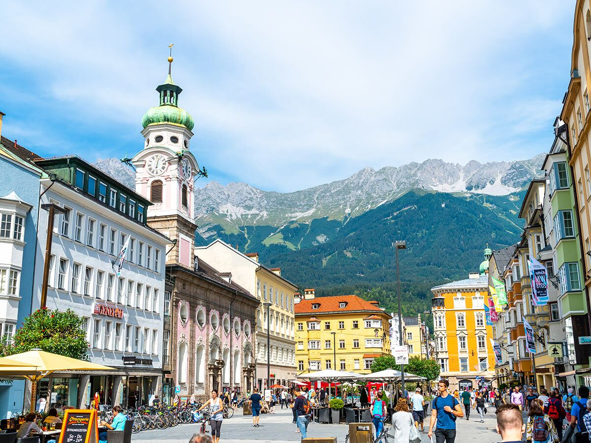 Belebter Marktplatz in Innsbruck mit historischem Glockenturm, bunten Häusern und majestätischen Alpen im Hintergrund bei Tag.
