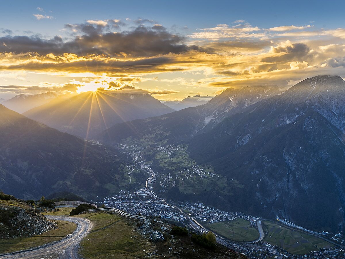Majestätische Alpenlandschaft bei Sonnenuntergang mit goldenen Sonnenstrahlen über einem Tal, das eine Stadt und einen Fluss umschließt; kurvige Bergstraße im Vordergrund.