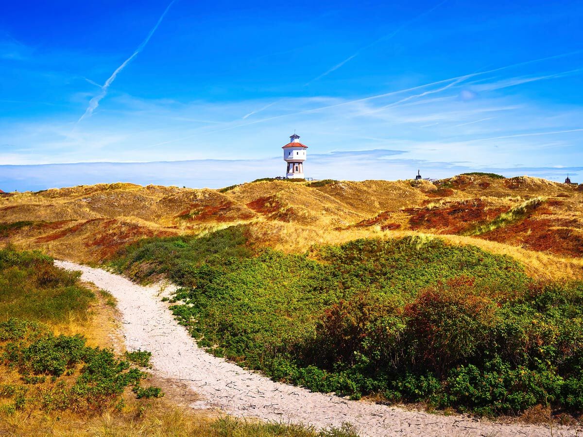 Weißer Wasserturm auf roten Dünen mit Sandpfad unter blauem Himmel, Nordsee-Küstenlandschaft.