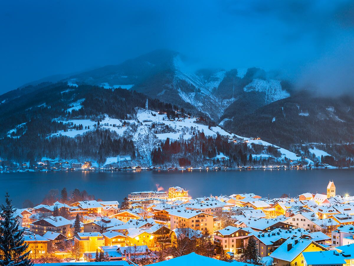 Blick auf das winterliche Zell am See in den österreichischen Alpen bei Nacht, mit beleuchteten Häusern, Schnee und einem See.