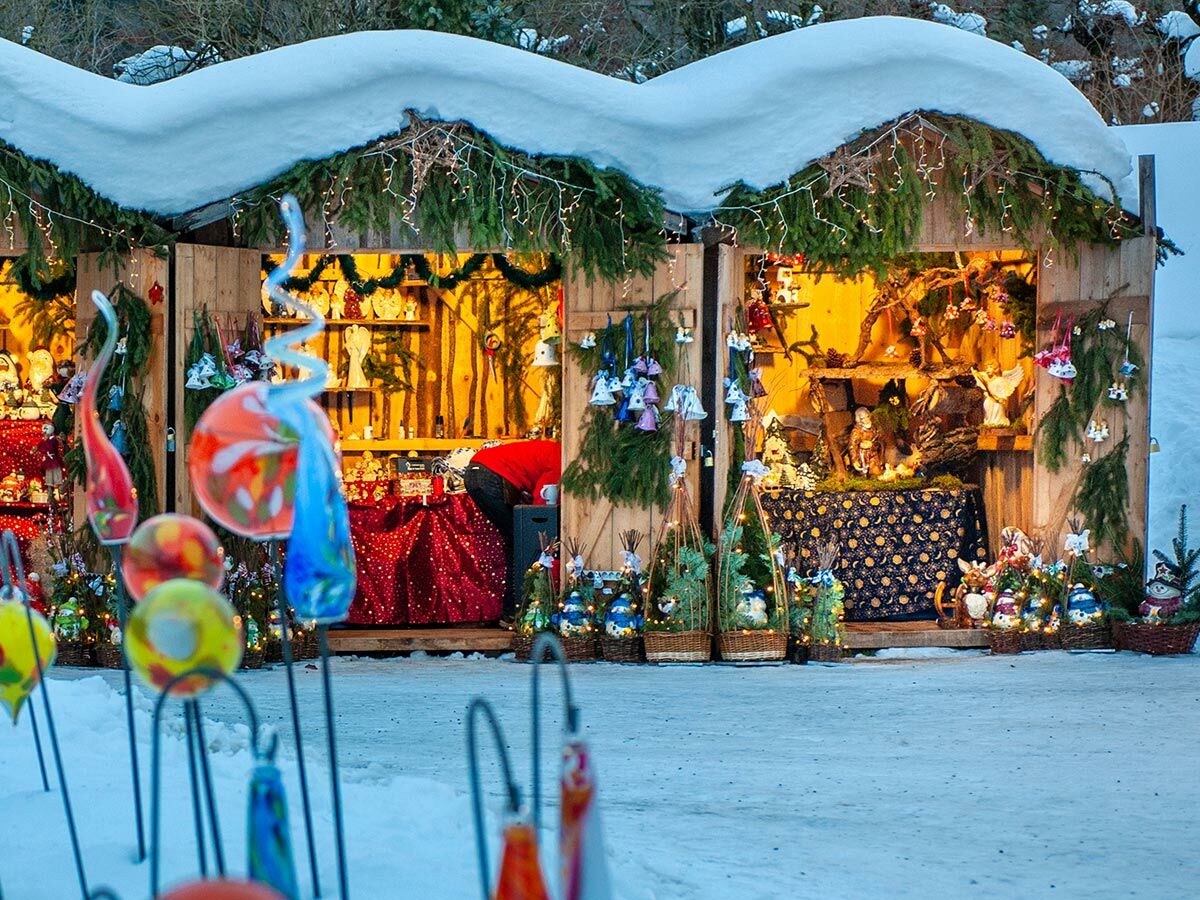 Festlich dekorierte Weihnachtsmarktstände mit Schneebedeckung, Lichtern, Christbaumschmuck und Kunsthandwerk im Schnee.