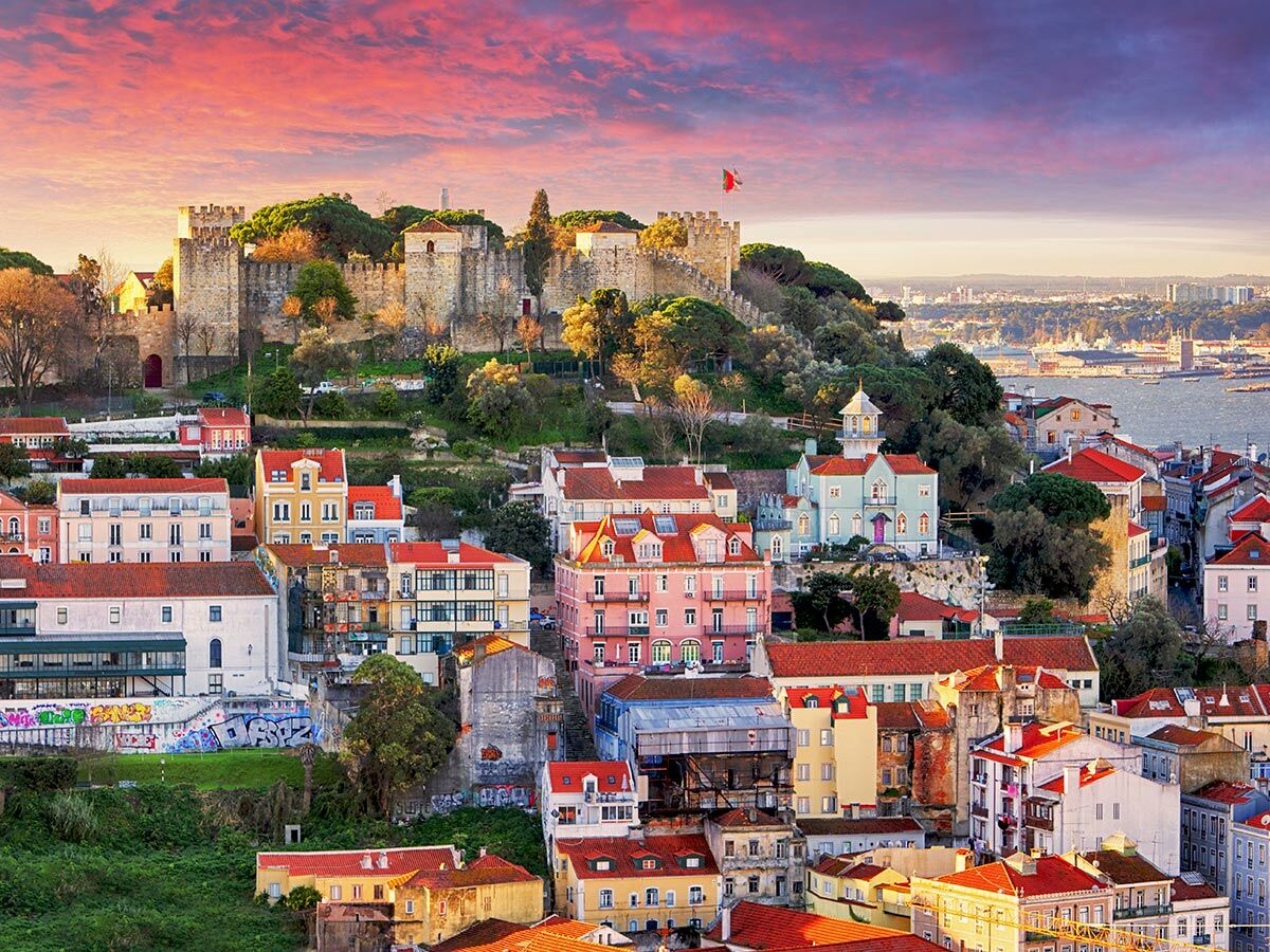Historische Burg São Jorge in Lissabon bei Sonnenuntergang. Bunte Altstadthäuser und der Tejo-Fluss unter rosa Himmel.
